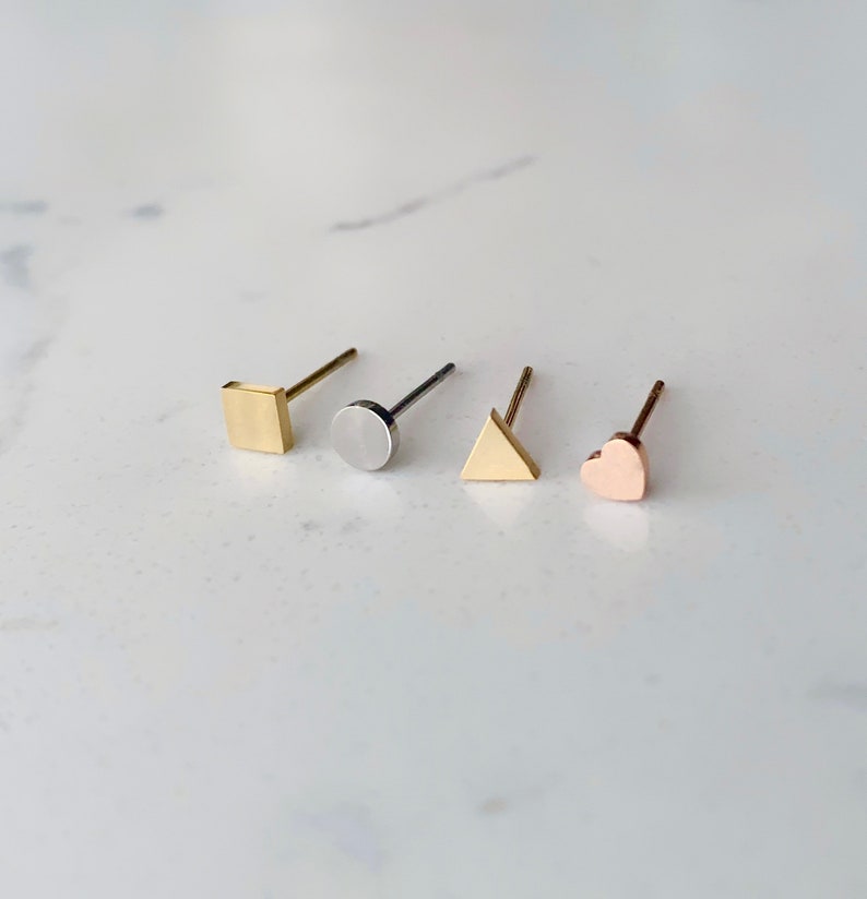 May include: Four gold stud earrings with different shapes: a square, a circle, a triangle, and a heart. The earrings are on a white marble surface.