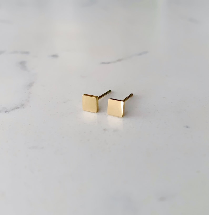 May include: A pair of gold square stud earrings. The earrings are small and simple, making them a versatile accessory for any occasion.