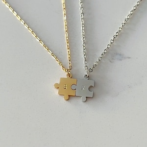 Op de afbeelding: Twee gouden en zilveren kettingen met puzzelstukjes als hangers. Het gouden puzzelstukje heeft de letter "B" erop gegraveerd en het zilveren puzzelstukje heeft de letter "R" erop gegraveerd.
