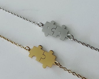 Peut inclure: Deux colliers en forme de pièce de puzzle, l'un en or et l'autre en argent. Les colliers sont sur un fond blanc.