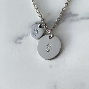 May include: Silver necklace with two charms. One charm is a small circle with a paw print. The other charm is a larger circle with the letter "S" engraved on it.