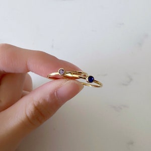 May include: Two gold rings with small gemstones. The ring on the left has a light pink gemstone, and the ring on the right has a dark blue gemstone.