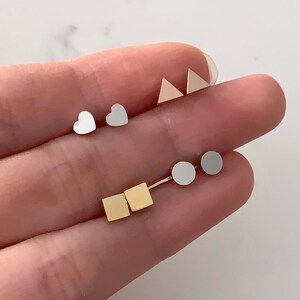 May include: A set of six small stud earrings in different shapes and colours. The earrings are silver, gold, and white. The shapes include a heart, a square, a circle, and a triangle.