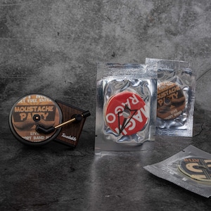 Peut inclure: Un tourne-disque miniature avec une base en bois brun et un bras argenté. Le tourne-disque joue un disque rouge et blanc avec le texte "RAGO" dessus. Il y a deux autres disques sur l'image, l'un avec le texte "Moustache Party" et l'autre avec le texte "Moustache Poty".