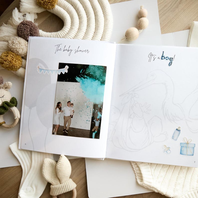 May include: Open baby shower memory book with the text "The baby shower" and "It's a boy!". The left page has a photo of a gender reveal with blue confetti. The right page has a stork illustration with a baby.