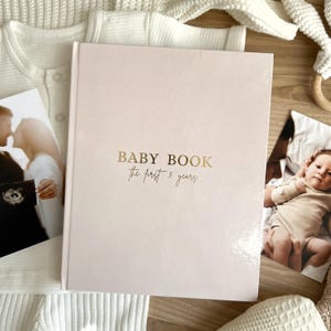 May include: A light pink baby book with gold lettering that reads "BABY BOOK the first 5 years." The book is open, with photos of a baby and parents on either side. A white onesie and a wooden toy are also in the image.