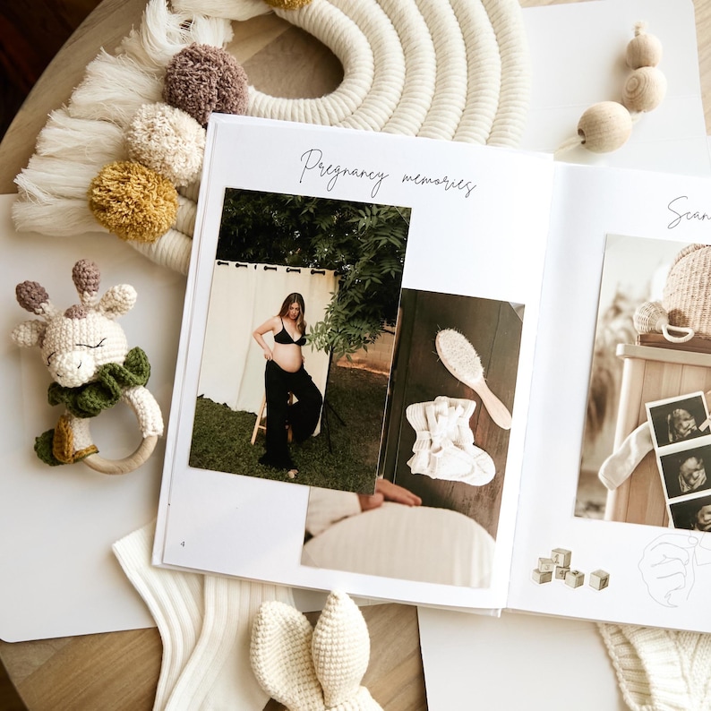 An open white photo album with the words "Pregnancy memories" in script. The album displays photographs, including a pregnant person in black clothing. A crocheted giraffe rattle, a macrame wall hanging, and other baby items are also visible.