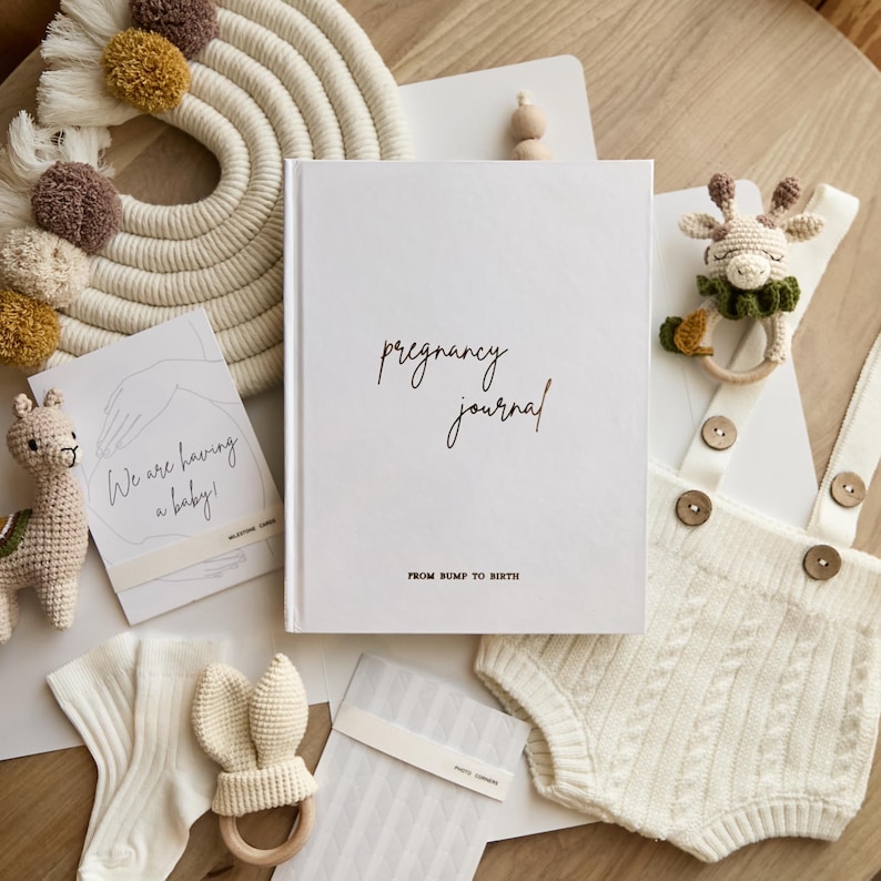 May include: A flat lay of baby items, including a white "pregnancy journal" book with the text "pregnancy journal" and "FROM BUMP TO BIRTH". Other items include a crochet giraffe rattle, a knitted romper, and a card that says "We are having a baby!"
