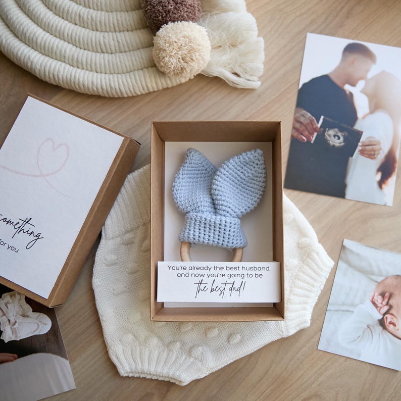 May include: A light blue crocheted baby teether with bunny ears attached to a wooden ring. The teether is in a brown box with a white card that reads "You're already the best husband and now you're going to be the best dad!"