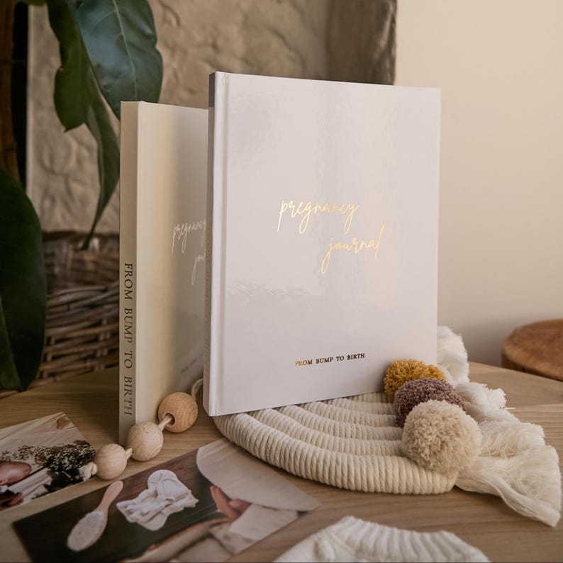 May include: Two hardcover pregnancy journals with gold lettering that reads "pregnancy journal" and "From Bump to Birth." The journals are white and cream-colored, resting on a woven mat with decorative pom-poms.