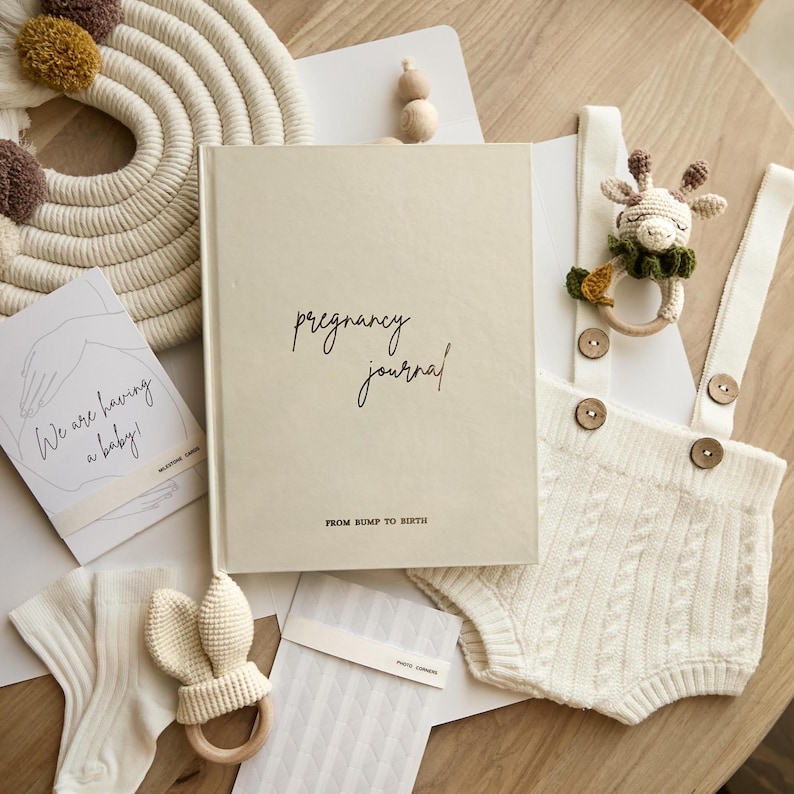 A cream-colored pregnancy journal with the words "pregnancy journal" and "FROM BUMP TO BIRTH" in black script. The image also includes baby clothes, a giraffe rattle, a card that says "We are having a baby!", and other baby accessories.