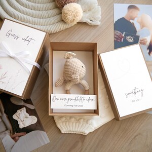 May include: A brown box with a white card that says "Guess what... One more grandchild to adore. Coming Fall 2025." Inside the box is a crocheted beige bunny rattle.