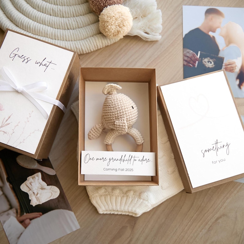 May include: A brown box with a white card that says "Guess what... One more grandchild to adore. Coming Fall 2025." Inside the box is a crocheted beige bunny rattle.