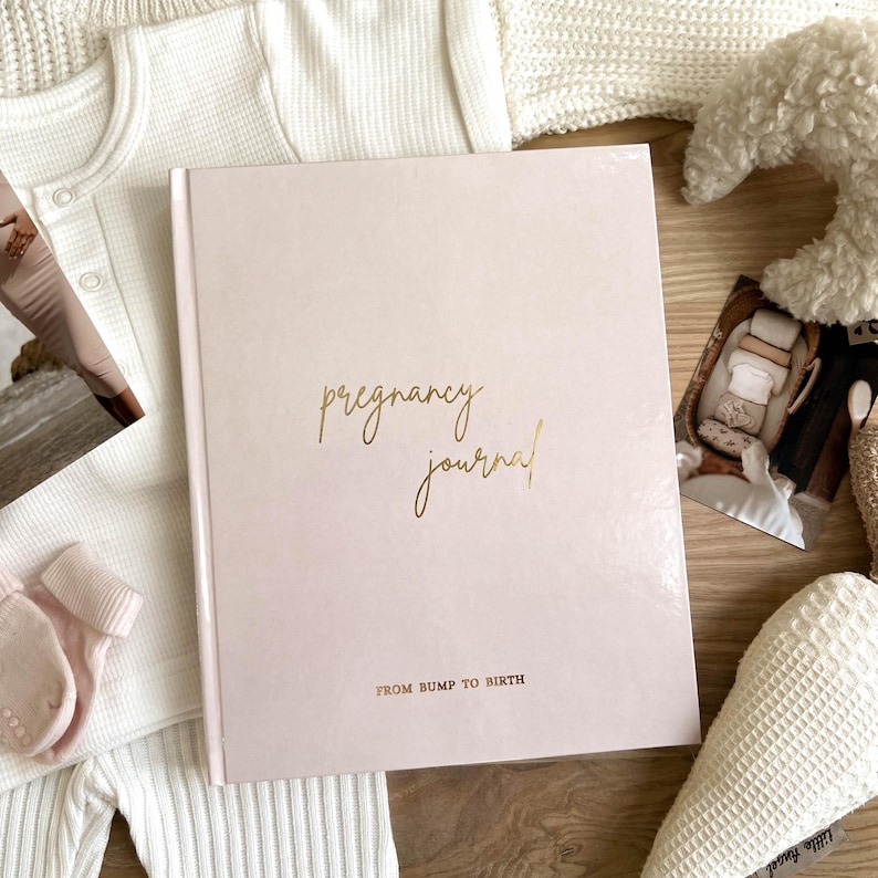 May include: A light pink pregnancy journal with gold lettering that reads "pregnancy journal" and "FROM BUMP TO BIRTH". The journal is surrounded by baby clothes, a photo, and a stuffed animal.