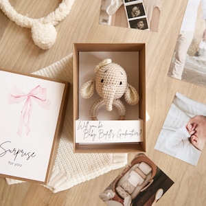 Puede incluir: Un sonajero de elefante de ganchillo beige en una caja marrón con el texto "Will you be Baby [Name]'s Godmother?" Una tarjeta de felicitación con un lazo rosa y la palabra "Surprise" está al lado de la caja. Fotos con temática de bebé en el fondo.