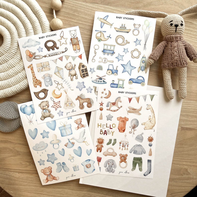 May include: Four sheets of baby-themed stickers with various illustrations, including teddy bears, stars, and vehicles. The stickers are in pastel colors and feature the text "BABY STICKERS" and "HELLO BABY". A crocheted bunny toy is also present.