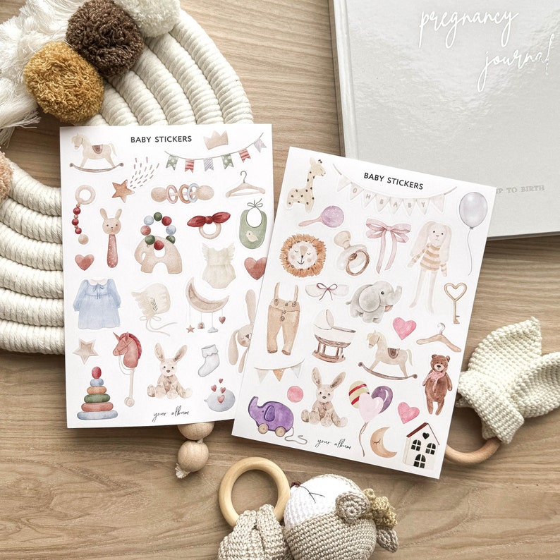 May include: Two sheets of baby stickers with watercolor illustrations of baby-related items like a rocking horse, bib, and rattle. The text "BABY STICKERS" is at the top of each sheet. A pregnancy journal is in the background.