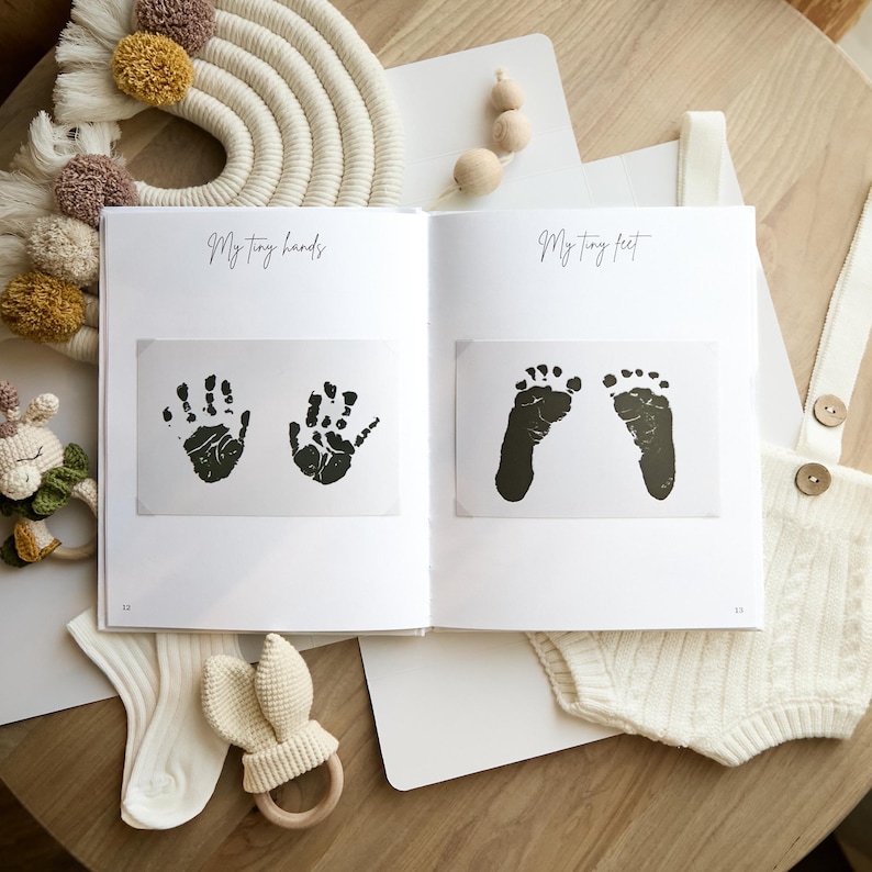May include: An open baby memory book with the text "My tiny hands" and "My tiny feet". The book displays black ink handprints and footprints. A white romper, socks, and a bunny-shaped wooden teether are visible.
