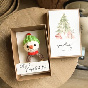 Puede incluir: Un sonajero de muñeco de nieve de ganchillo con gorro verde, bufanda roja y brazos marrones, presentado en una caja marrón con una tarjeta que dice "Will you be Mary's Godmother?" y una tarjeta con una ilustración de árbol de Navidad y "something for you."