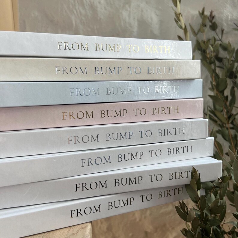 May include: A stack of books with the title "FROM BUMP TO BIRTH" in gold lettering. The books are in various pastel colors, including white, beige, light blue, and pink. The books are stacked on top of each other.