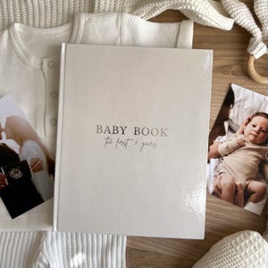 Op de afbeelding: Een crèmekleurig babyboek met harde kaft en de tekst "BABY BOOK the first 5 years" in zilver op de voorkant gedrukt. Het boek is omringd door een wit kledingstuk met wafelstructuur en twee foto's van een baby en een echografie.