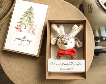 Puede incluir: Una caja de regalo de cartón marrón que contiene un pequeño reno de ganchillo con una pajarita roja. La tapa presenta una ilustración en acuarela de un árbol de Navidad y el texto "something for you". Una tarjeta interior dice "One more grandchild to adore... Coming 2027".