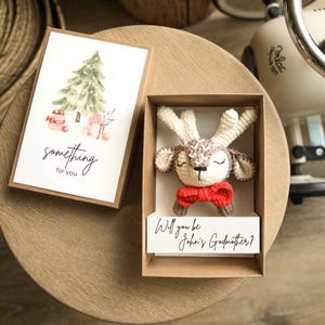 Puede incluir: Un reno de ganchillo con pajarita roja se presenta en una caja de regalo. Una tarjeta con una ilustración de árbol de Navidad y el texto "something for you" está al lado de la caja. El texto "Will you be John's Godmother?" está en una tarjeta.
