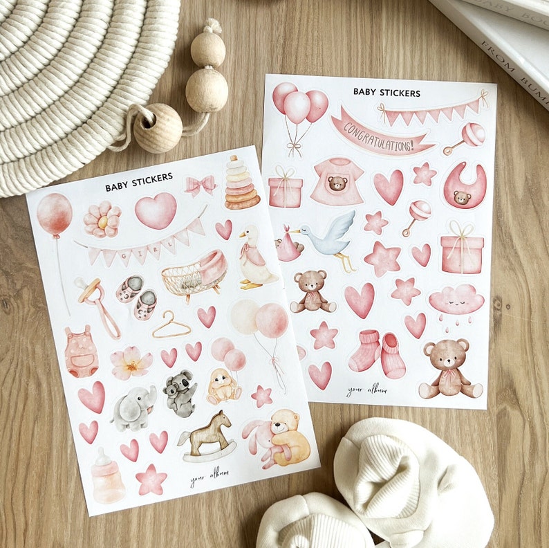 May include: Two sheets of baby stickers with various pink illustrations, including balloons, teddy bears, hearts, and baby clothes. The text "BABY STICKERS" and "CONGRATULATIONS!" are visible. The stickers are on a wooden surface.