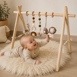 Könnte beinhalten: Ein hölzernes Spieltrapez für Babys mit fünf hängenden Häkelspielzeugen, darunter ein Lama, ein Regenbogen, eine Perlenkette, eine Eule und ein Löwe. Die Spielzeuge sind an Holzringen an einer horizontalen Stange befestigt.