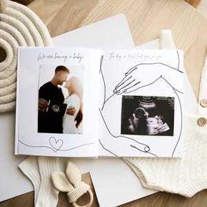 Open photo album with the text "We are having a baby!" and "The day we first laid eyes on you." The album features a photo of a couple and an ultrasound image. A bunny-shaped teether and baby clothes are also visible.