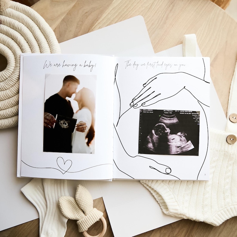 Open photo album with the text "We are having a baby!" and "The day we first laid eyes on you." The album features a photo of a couple and an ultrasound image. A bunny-shaped teether and baby clothes are also visible.