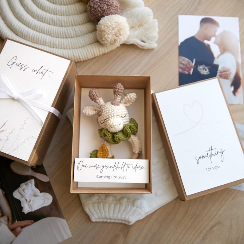May include: A brown box with a white card that says "Guess what" and a white ribbon. Inside the box is a crocheted giraffe rattle with green leaves and a brown and yellow leaf. The card inside the box says "One more grandchild to adore, Coming Fall 2025".