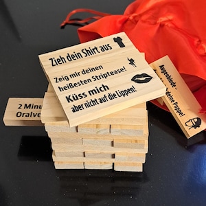 Może przedstawiać: Drewniana gra blokowa z nadrukowanymi frazami i ilustracjami. Klocki są ułożone w stos, niektóre z tekstem, takim jak "Zdejmij koszulę" i "Pocałuj mnie". Widoczny jest również pomarańczowy worek ze sznurkiem i mały drewniany element z tekstem.