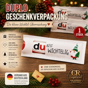 Op de afbeelding: Een witte rechthoekige papieren hoes voor een chocoladereep met de tekst 'DU BIST WICHTELIG' en een afbeelding van een kabouter die een klein boompje vasthoudt. De hoes heeft een uitgesneden venster en meet 14 bij 5 cm.
