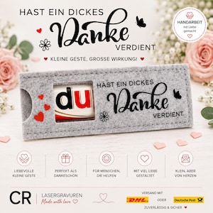 Op de afbeelding: Een rechthoekige hoes van grijs vilt met een uitgesneden venster dat een chocoladereep onthult. De hoes bevat zwarte tekst HAST EIN DICKES DANKE VERDIENT, een kleine zwarte vlinder, een bloemicoon en drie kleine rode hartjes aan de linkerkant.