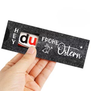 Könnte beinhalten: Ein dunkelgrauer Oster-Geschenkehalter aus Filz mit den Worten "HEY DU FROHE OSTERN" in Weiß. Der Halter hat ein rechteckiges Fenster, das einen Teil eines Schokoriegels zeigt. Eine Hand hält den Geschenkehalter.