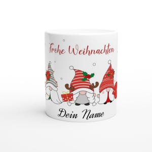 Op de afbeelding: Witte keramische mok met drie kerstkabouters met rode en witte hoeden. De mok heeft de tekst "Frohe Weihnachten" en "Dein Name" in het rood. De kabouters houden cadeaus vast en zijn feestelijk gekleed.