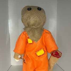 May include: A handmade Halloween doll with an orange jumpsuit and yellow patches, a burlap sack head with button eyes, and a beige glove holding a candy. The doll is holding a beige sack with a red and brown top.