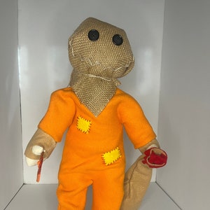 May include: A handmade plush doll of Sam from the horror film Trick 'r Treat. The doll has a burlap sack head with button eyes, an orange jumpsuit with yellow patches, and a brown sack. It holds a candy cane.