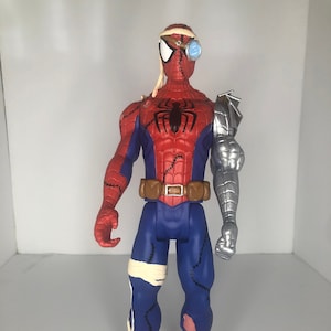 May include: A Spider-Man action figure with a unique design. The figure features a red and blue suit with stitched details, a brown belt, and a silver robotic arm. It has a bandage on its head and leg, and a steampunk-style eye patch.