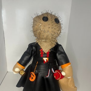 May include: A handmade doll with a burlap sack head, secured with pins, and button eyes. It wears a black leather-like outfit with orange accents, a belt, and a small pumpkin-shaped accessory. The doll has tan fabric hands and orange and white feet.