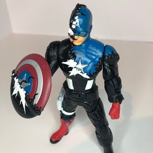 Puede incluir: Figura de acci&oacute;n de Capit&aacute;n Am&eacute;rica con una combinaci&oacute;n de colores azul y negro. La figura sostiene un escudo rojo, blanco y azul. El dise&ntilde;o incorpora una est&eacute;tica similar a la de un simbionte, con zarcillos negros y un efecto de color dividido.