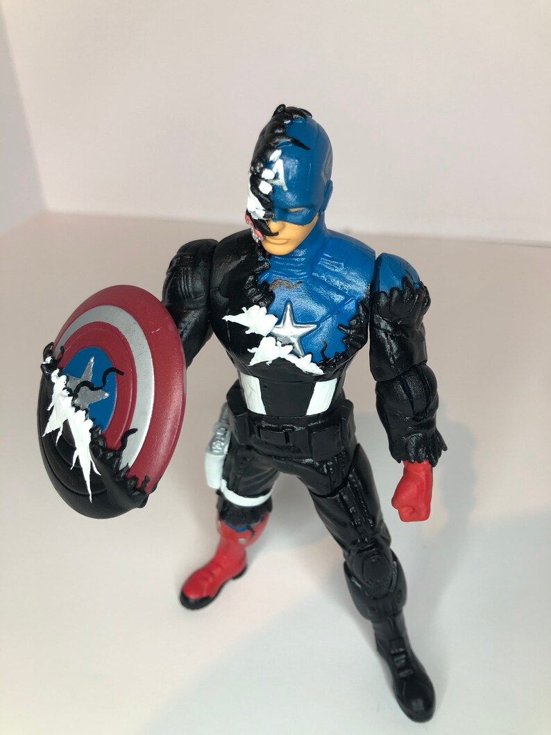 Puede incluir: Figura de acci&oacute;n de Capit&aacute;n Am&eacute;rica con una combinaci&oacute;n de colores azul y negro. La figura sostiene un escudo rojo, blanco y azul. El dise&ntilde;o incorpora una est&eacute;tica similar a la de un simbionte, con zarcillos negros y un efecto de color dividido.