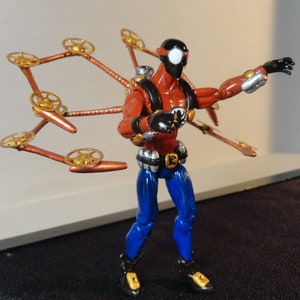 May include: A red and blue action figure with a black mask and steampunk-style wings. The figure has a black and white spider-like mask, red torso, blue pants, and metallic accents. The wings are copper-colored with gold gears.