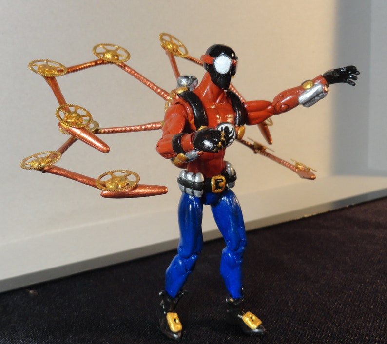 May include: A red and blue action figure with a black mask and steampunk-style wings. The figure has a black and white spider-like mask, red torso, blue pants, and metallic accents. The wings are copper-colored with gold gears.