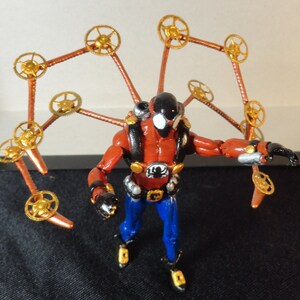 May include: A red and black action figure with blue pants and gold accents. The figure has a black helmet and a backpack. Attached to the figure are copper-colored arms with gold gears at the ends, resembling wings.
