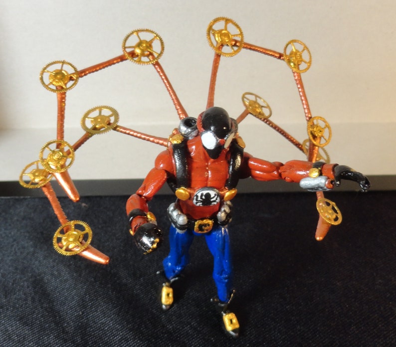 May include: A red and black action figure with blue pants and gold accents. The figure has a black helmet and a backpack. Attached to the figure are copper-colored arms with gold gears at the ends, resembling wings.