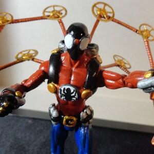 May include: A red and black action figure with mechanical spider-like arms. The figure has a black mask, a spider emblem on the chest, and blue pants. The arms are copper-colored with gold watch gears.