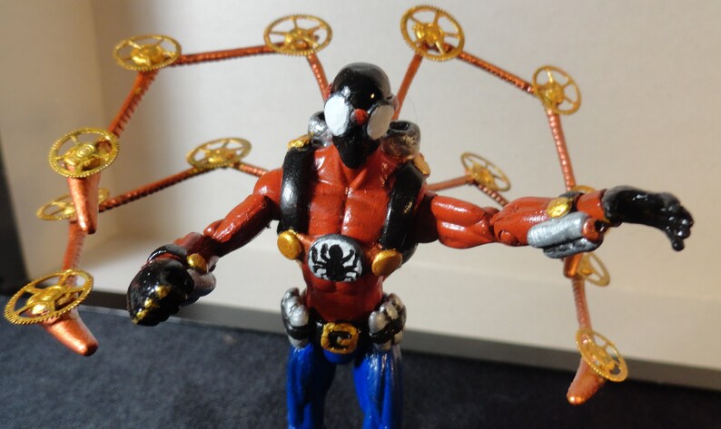May include: A red and black action figure with mechanical spider-like arms. The figure has a black mask, a spider emblem on the chest, and blue pants. The arms are copper-colored with gold watch gears.