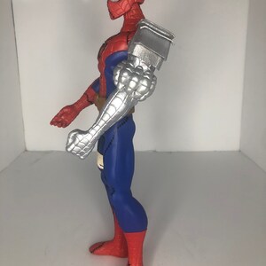 May include: A Spider-Man action figure with a steampunk-inspired design. The figure features a red and blue suit, a silver robotic arm, and goggles. The figure is approximately 12 inches tall.